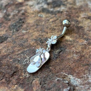 May include: A silver belly button ring featuring a flip-flop charm with clear accents. The charm is suspended from a curved barbell with a small ball at the top. The flip-flop design includes a flower and additional clear embellishments.