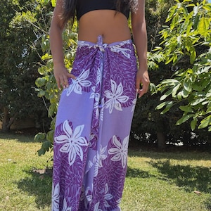 May include: A purple and white floral sarong tied at the waist. The sarong features a large white flower and leaf pattern on a purple background. The person is standing on green grass in front of a tree.