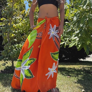 May include: An orange sarong with a floral design featuring white flowers and green leaves. The sarong is tied at the waist and worn over a black tube top. The background includes green trees and grass.