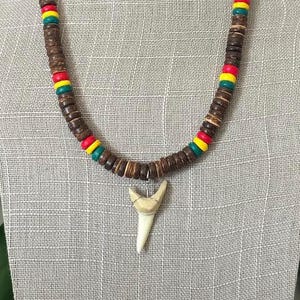 May include: A brown beaded necklace with red, yellow, and green accents. The necklace features a pendant shaped like a shark tooth. The necklace is displayed on a gray mannequin.