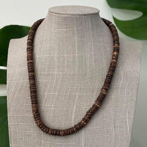 May include: A dark brown beaded necklace displayed on a mannequin. The necklace is made of round, wooden beads. The beads are strung together to form a simple, yet elegant necklace. The necklace is a natural, earthy accessory.