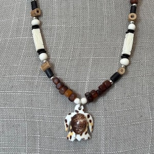 May include: A beaded necklace featuring a turtle pendant. The necklace has a mix of brown, black, white, and silver beads. The turtle pendant is white with brown spots and a brown shell. The necklace is displayed on a mannequin.