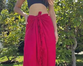 May include: A woman's outfit featuring a dark blue tube top and a long, vibrant red sarong. The sarong is tied at the waist and has a decorative fringe detail along the side. The outfit is worn outdoors in front of green foliage.