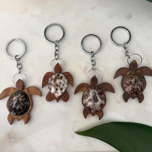 May include: Four wooden turtle keychains with shell accents. Each keychain features a turtle shape with a natural wood finish and a shell center. The keychains have silver-colored key rings and chains.
