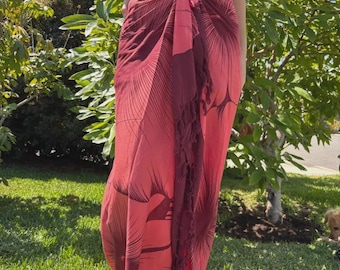 May include: A red and maroon sarong with a palm leaf design. The sarong is tied at the waist and has a fringed edge. The sarong is worn outdoors on a sunny day.