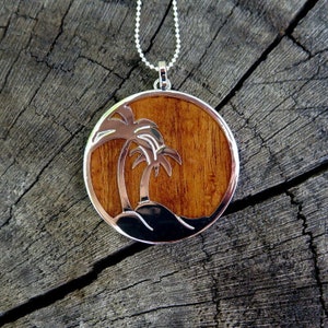 May include: A silver necklace with a round pendant featuring a woodgrain design and two palm trees cut out of the metal. The pendant is hanging on a silver chain.