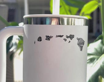 Puede incluir: Taza de viaje blanca con borde y asa plateados. La taza presenta un gráfico negro de las islas hawaianas. Una pajita blanca se extiende desde la tapa. La taza está al aire libre con follaje verde en el fondo.