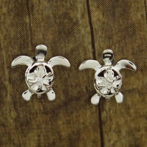 May include: A pair of silver-colored turtle stud earrings. Each earring features a turtle design with a flower detail in the center of the shell. The earrings are displayed on a brown, textured surface.