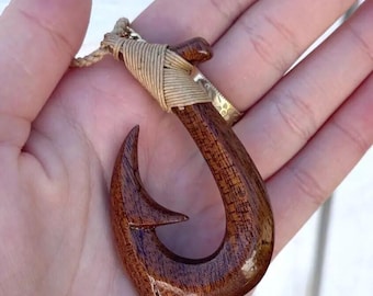 Puede incluir: Colgante artesanal de anzuelo de pesca de madera con acabado natural. El anzuelo es de color marrón oscuro, con un cordón tejido y detalles metálicos dorados. El colgante se sostiene en una mano.