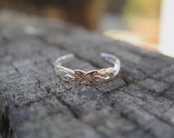 May include: A silver-toned adjustable ring with a floral design. The ring features a central flower in a rose gold hue, flanked by detailed silver patterns. The ring is set against a textured, gray wooden surface.