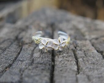 May include: A silver-toned ring with two white flower designs, each with a small, clear gem in the center. The ring has a gold-colored band with an etched pattern. The ring is set against a weathered, gray wooden surface.