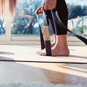 May include: A black yoga strap with a wooden accent is held by a person wearing black leggings. The strap is positioned on a light-colored yoga mat. The background includes a window with a view of trees and a bright sky.