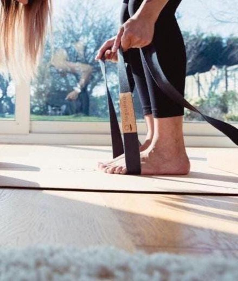 May include: A black yoga strap with a wooden accent is held by a person wearing black leggings. The strap is positioned on a light-colored yoga mat. The background includes a window with a view of trees and a bright sky.