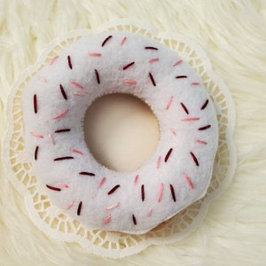 May include: White felt donut with pink and burgundy sprinkles. The donut is on a white doily.