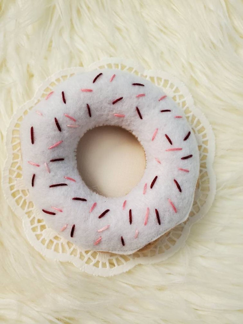 May include: White felt donut with pink and burgundy sprinkles. The donut is on a white doily.