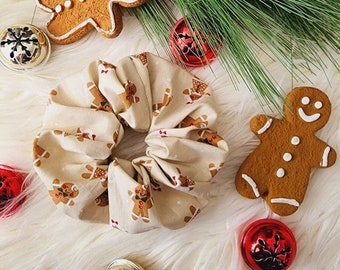 May include: A beige scrunchie with a gingerbread man pattern. The scrunchie is surrounded by gingerbread men cookies and red and gold bells on a white fluffy surface.