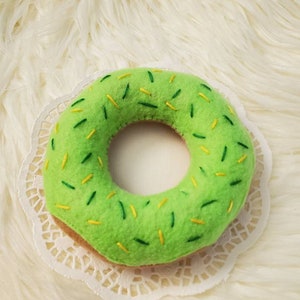 May include: A green felt donut with yellow and green sprinkles. The donut is on a white doily.