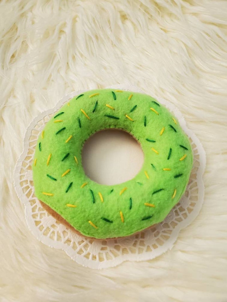 May include: A green felt donut with yellow and green sprinkles. The donut is on a white doily.