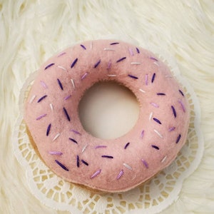 May include: A pink felt donut with purple and white sprinkles. The donut is sitting on a white doily.