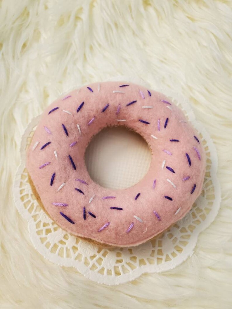 May include: A pink felt donut with purple and white sprinkles. The donut is sitting on a white doily.