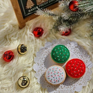 May include: Three felt Christmas cookies with white frosting and colorful sprinkles. The cookies are on a white doily on a white fluffy surface. There are red and gold ornaments in the background.