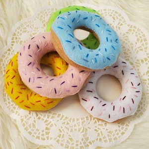 May include: Four felt donuts with colorful sprinkles. The donuts are yellow, pink, blue, and white. They are arranged on a white doily.