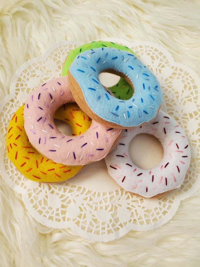 May include: Four felt donuts with colorful sprinkles. The donuts are yellow, pink, blue, and white. They are arranged on a white doily.