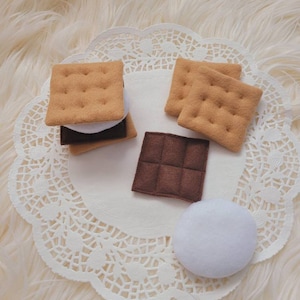 Peut inclure: Un ensemble d'aliments en feutre pour jouer à faire semblant, comprenant deux biscuits graham, une barre de chocolat et une guimauve. Les articles sont disposés sur une dentelle blanche.