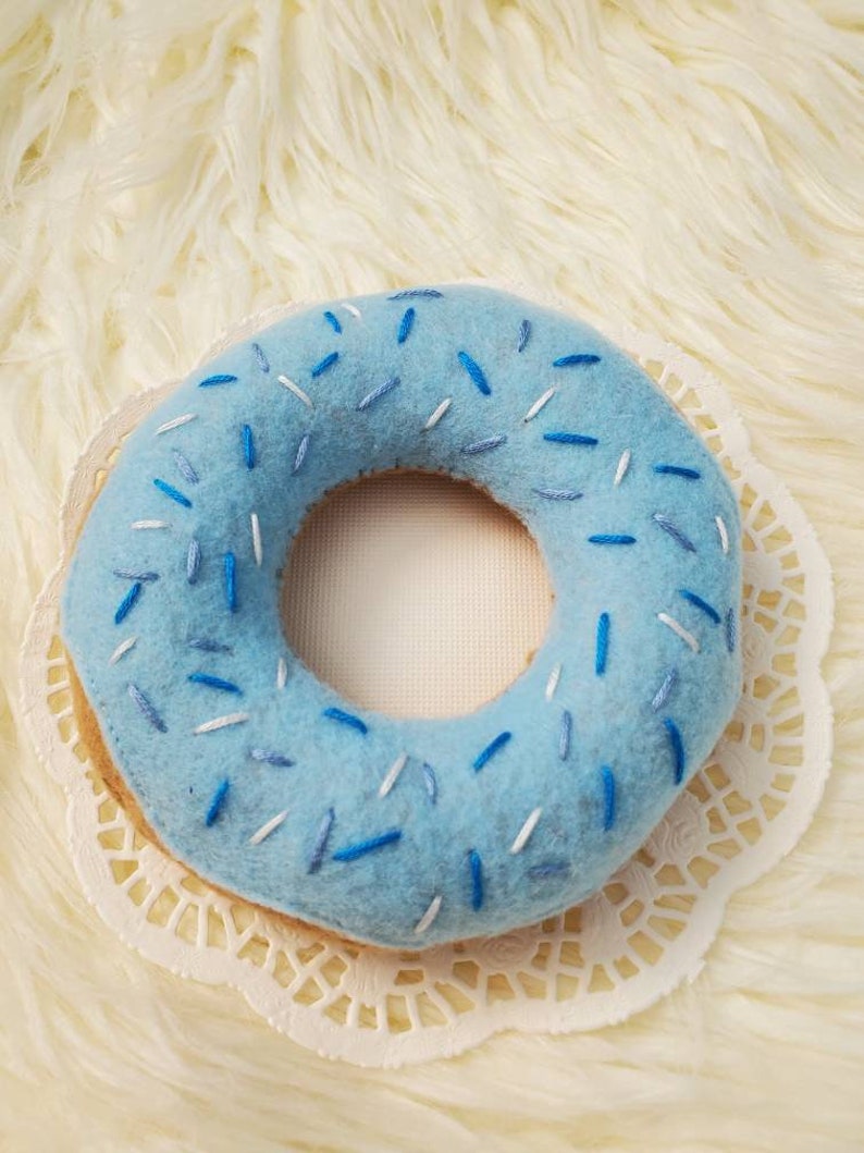 May include: A blue felt donut with white and blue sprinkles. The donut is on a white doily.