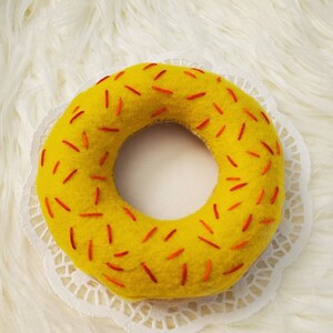 May include: A yellow felt donut with red sprinkles. The donut is on a white doily.