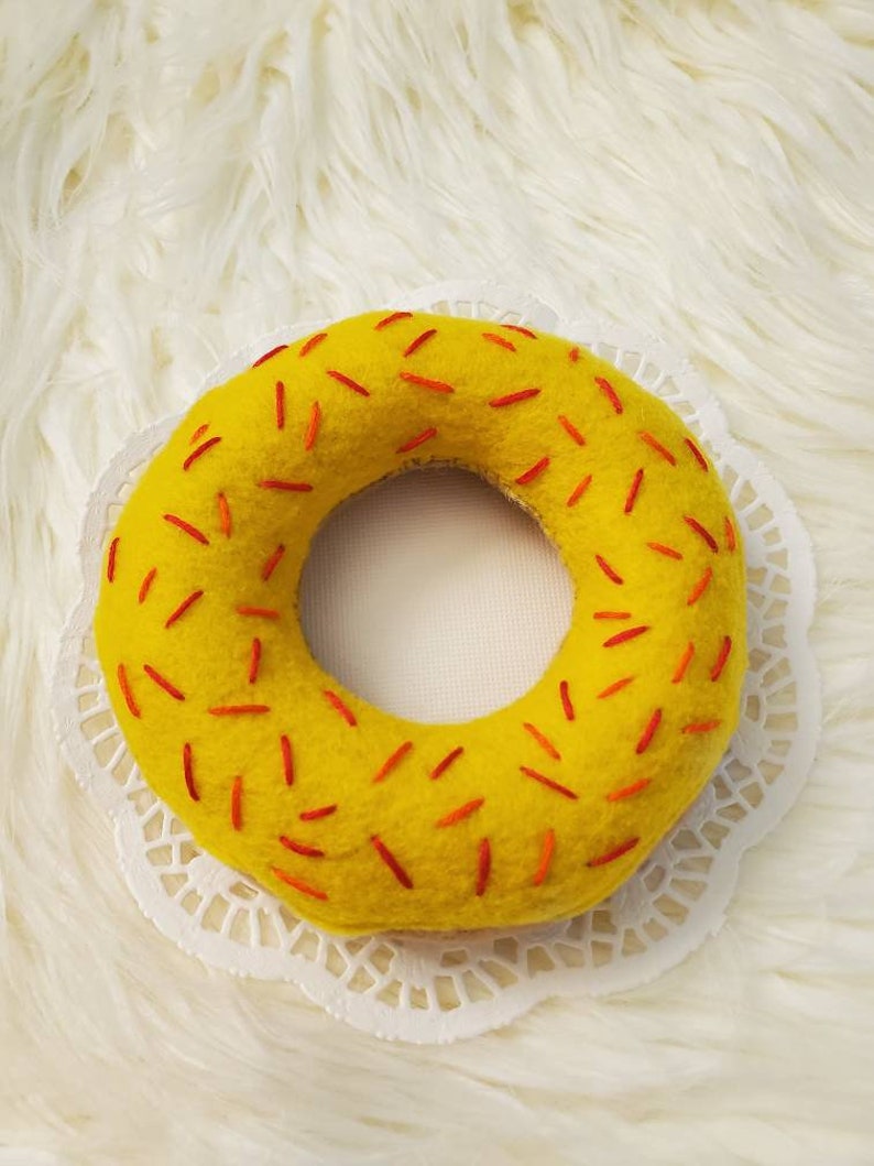 May include: A yellow felt donut with red sprinkles. The donut is on a white doily.
