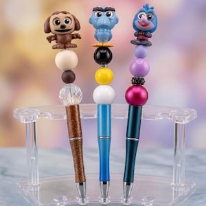 May include: Three decorative pens with character toppers. The pens are displayed in a clear plastic holder. The first pen has a brown dog topper, the second has a blue bird-like character, and the third has a blue bird-like character. Each pen has a unique bead design.