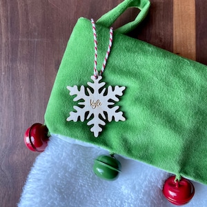 May include: Green and white Christmas stocking with a snowflake ornament that says "Kyle" hanging from it. The stocking has red and green bells.