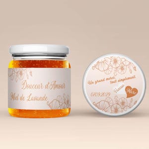 Peut inclure: Un pot rond de miel avec un couvercle blanc et une étiquette qui indique "Douceur d'Amour Miel de Lavande". L'étiquette a un motif floral et est de couleur rose clair. Un autocollant rond avec un motif floral et le texte "Un grand merci, tout simplement. 07.09.2029 Vanessa Marc" est également présenté.