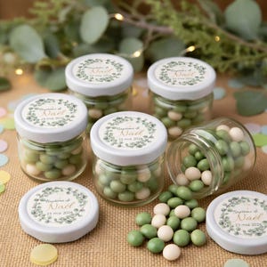 Peut inclure: Plusieurs petits pots en verre remplis de bonbons verts et blancs, dont certains renversés. Les pots ont des couvercles blancs avec une étiquette indiquant "Baptême de Noël 23 mai 2026" dans un motif de couronne. Le fond comprend des feuilles.