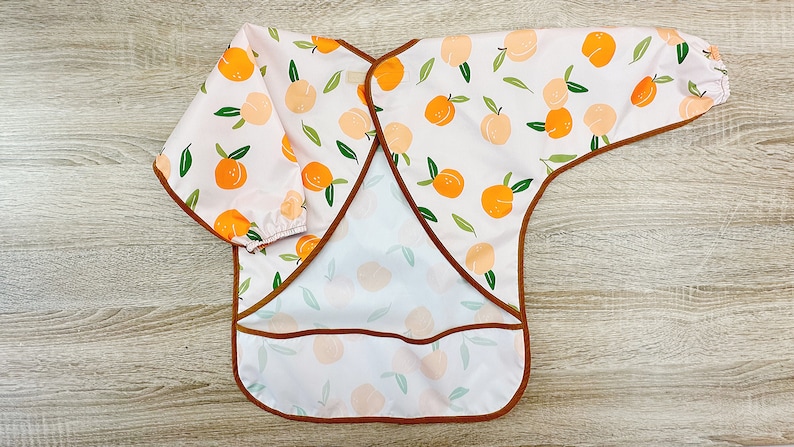 K&ouml;nnte beinhalten: Eine rosa-wei&szlig;e, lang&auml;rmelige L&auml;tzchen mit einem Pfirsichmuster und braunem Besatz. Das L&auml;tzchen hat eine Tasche auf der Vorderseite.