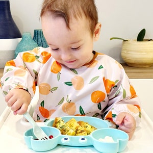 K&ouml;nnte beinhalten: Ein Baby, das eine L&auml;tzchen mit Pfirsichmuster tr&auml;gt, sitzt in einem Hochstuhl und isst von einem blauen Silikonteller in Form einer Krabbe. Der Teller hat drei F&auml;cher und das Baby benutzt eine blaue und wei&szlig;e Plastikgabel zum Essen.