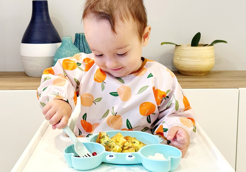 K&ouml;nnte beinhalten: Ein Baby, das eine L&auml;tzchen mit Pfirsichmuster tr&auml;gt, sitzt in einem Hochstuhl und isst von einem blauen Silikonteller in Form einer Krabbe. Der Teller hat drei F&auml;cher und das Baby benutzt eine blaue und wei&szlig;e Plastikgabel zum Essen.