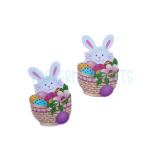 May include: Two white bunny rabbit figurines holding baskets filled with colorful Easter eggs. The baskets are woven and have pink ribbon accents. The bunnies have pink ears and noses.