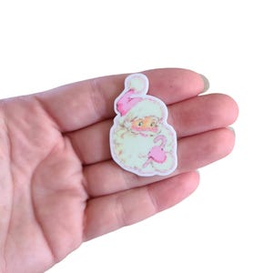 May include: A white and pink Christmas ornament featuring a vintage illustration of Santa Claus holding a candy cane.