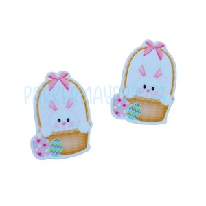 May include: Two white bunny rabbit figurines in a brown basket with pink bows and colorful Easter eggs.