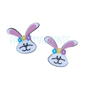 May include: A pair of white bunny rabbit earrings with pink ears and a flower crown. The flowers are yellow, blue, and purple.