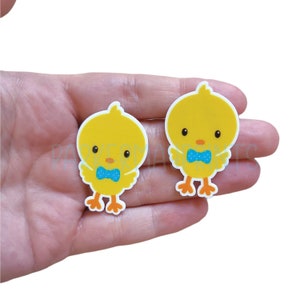 May include: Two yellow chick cartoon cutouts with blue bow ties. The chicks have orange feet and beaks. The cutouts are on a hand.