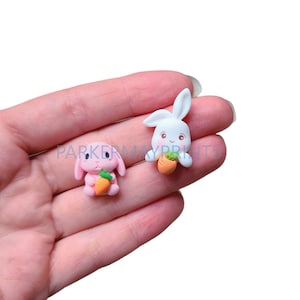 May include: Two small, handmade polymer clay bunny figurines. One bunny is pink and the other is white. Both bunnies are holding a small orange carrot.