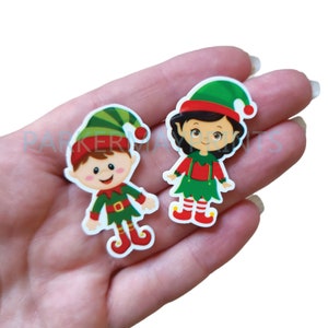 May include: Two Christmas elf figurines, one male and one female, with green hats, red and green outfits, and white boots. The figurines are on a white background.