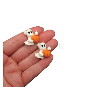 May include: Two white ghost figurines holding orange pumpkins. The ghosts have black eyes and a small smile. The pumpkins are a bright orange color.