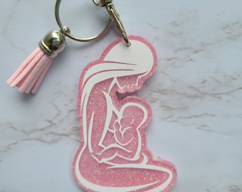 May include: Pink glitter keychain with a white silhouette of a mother holding a baby. The keychain has a silver tassel.