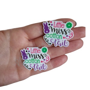 May include: Two white, round, resin embellishments with the text "Little miss cotton tail" in pink, green, purple, and black lettering. The embellishments feature a bunny rabbit with purple ears and a green flower.