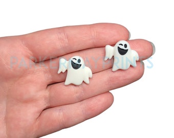 May include: Two white ghost-shaped charms with black eyes and smiling mouths. The ghosts are winking and have a white outline.
