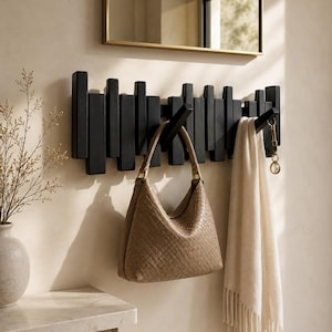 May include: A wall-mounted coat rack featuring a series of vertical black wooden slats of varying heights. The rack includes several angled hooks, with a tan woven handbag hanging from one and a cream-colored fringed scarf from another.